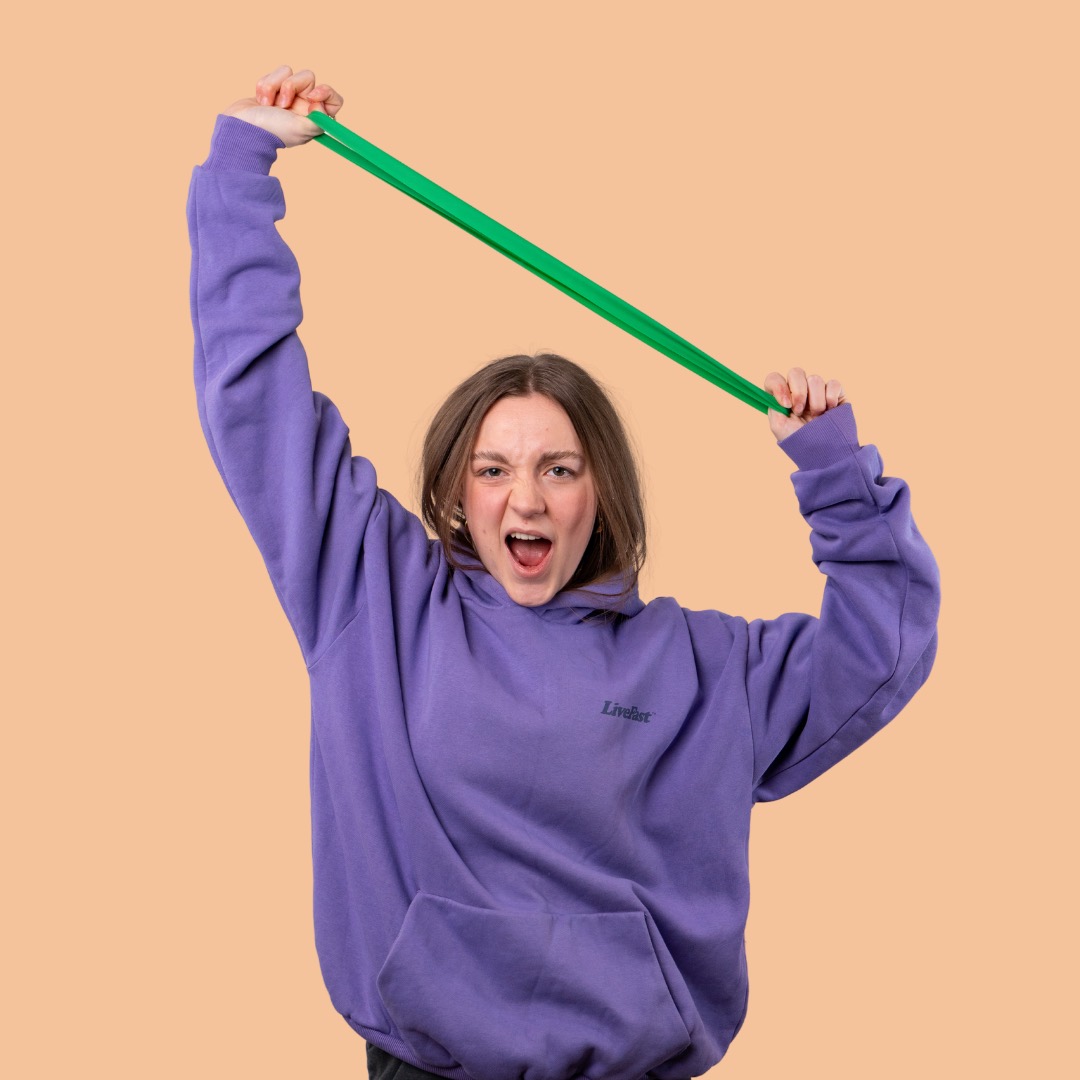Frau im lila Hoodie streckt ein Fitnessband über den Kopf