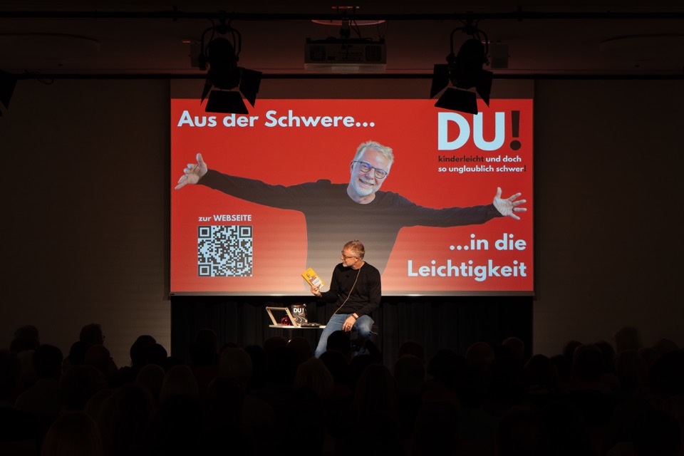 Redner auf dem Barhocker vor der großen Leinwand im abgedunkelten Saal